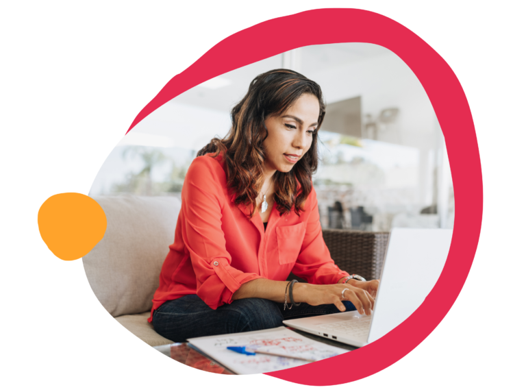 working woman in red shirt using laptop, focused expression, casual setting with notebook and pen on table, remote work lifestyle