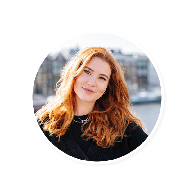 smiling woman with wavy red hair wearing a black sweater, sunlit cityscape background suggesting a cheerful atmosphere