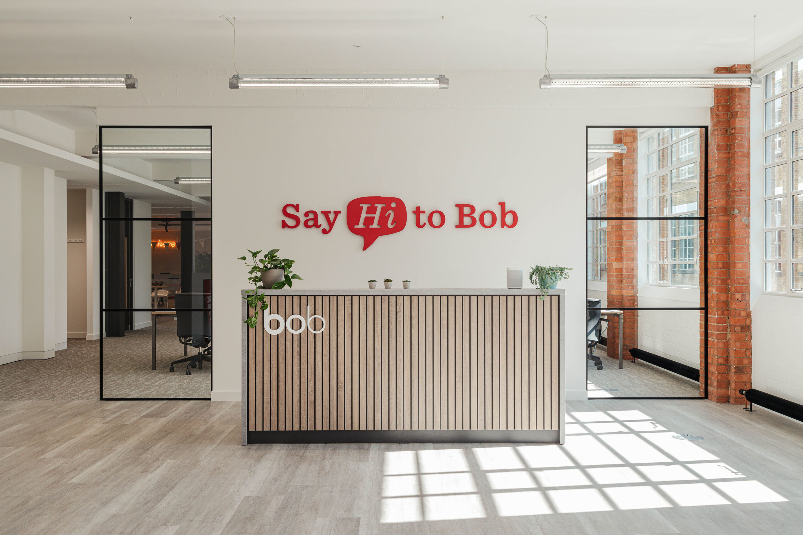 Say Hi to Bob, Modern office reception with wooden desk and potted plants, bright natural light through large windows.