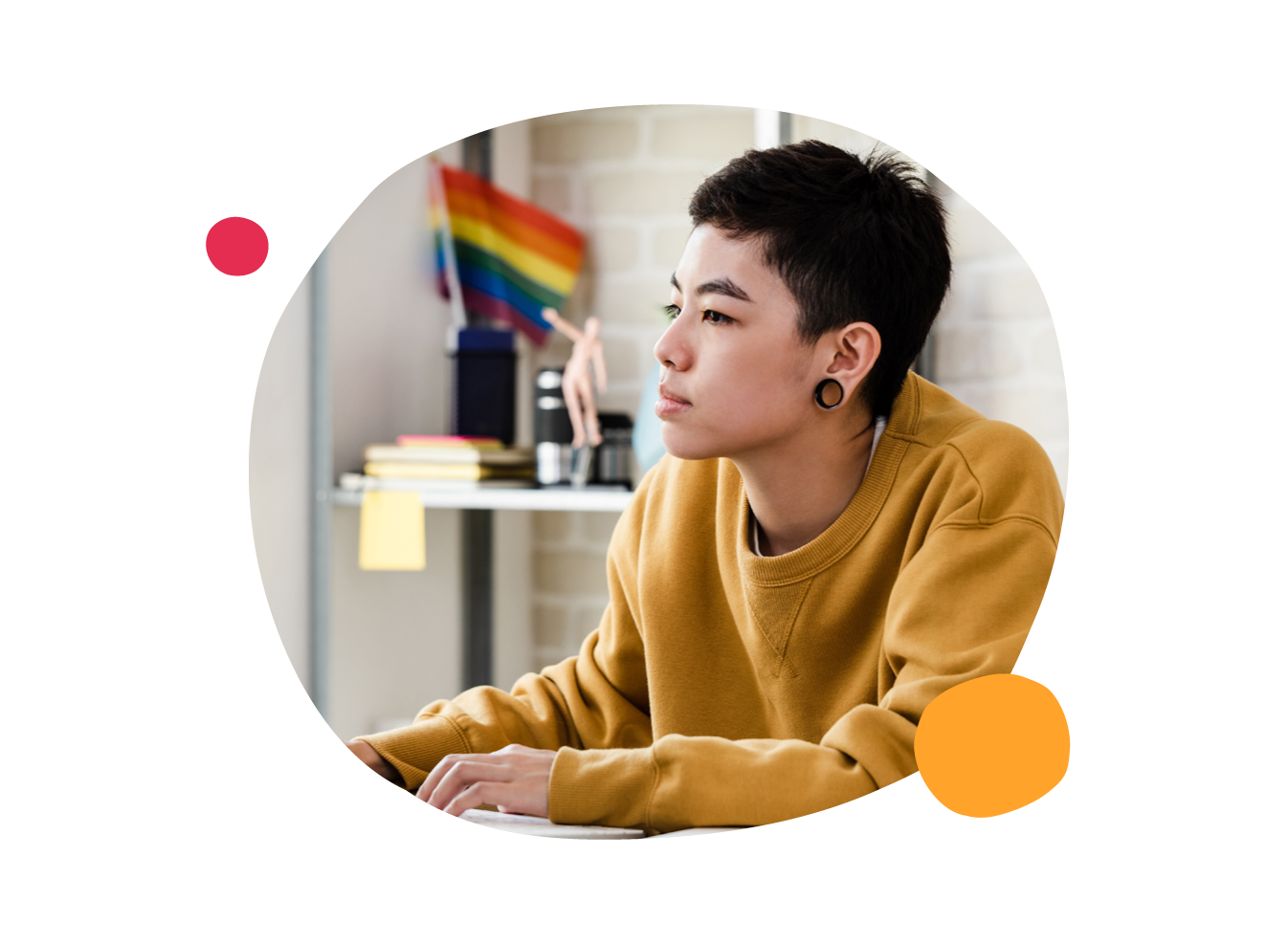 young person with earrings in a yellow sweater, focused at a desk with colorful books and a rainbow flag in the background, creative workspace inspiration