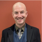 man in suit smiling against red background, professional attire with plaid tie and bald head