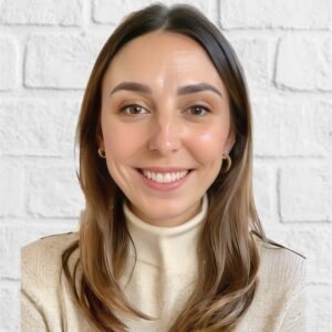 smiling woman with long brown hair in a cream sweater against a textured white brick wall, portrait with warm expressions and neutral tones
